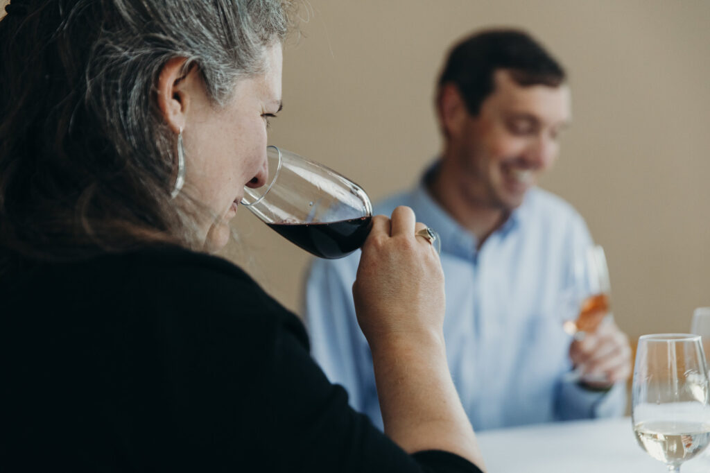 woman holding glass of red wine to her nose to smell aroma. Blurry image of a man is seen in background.