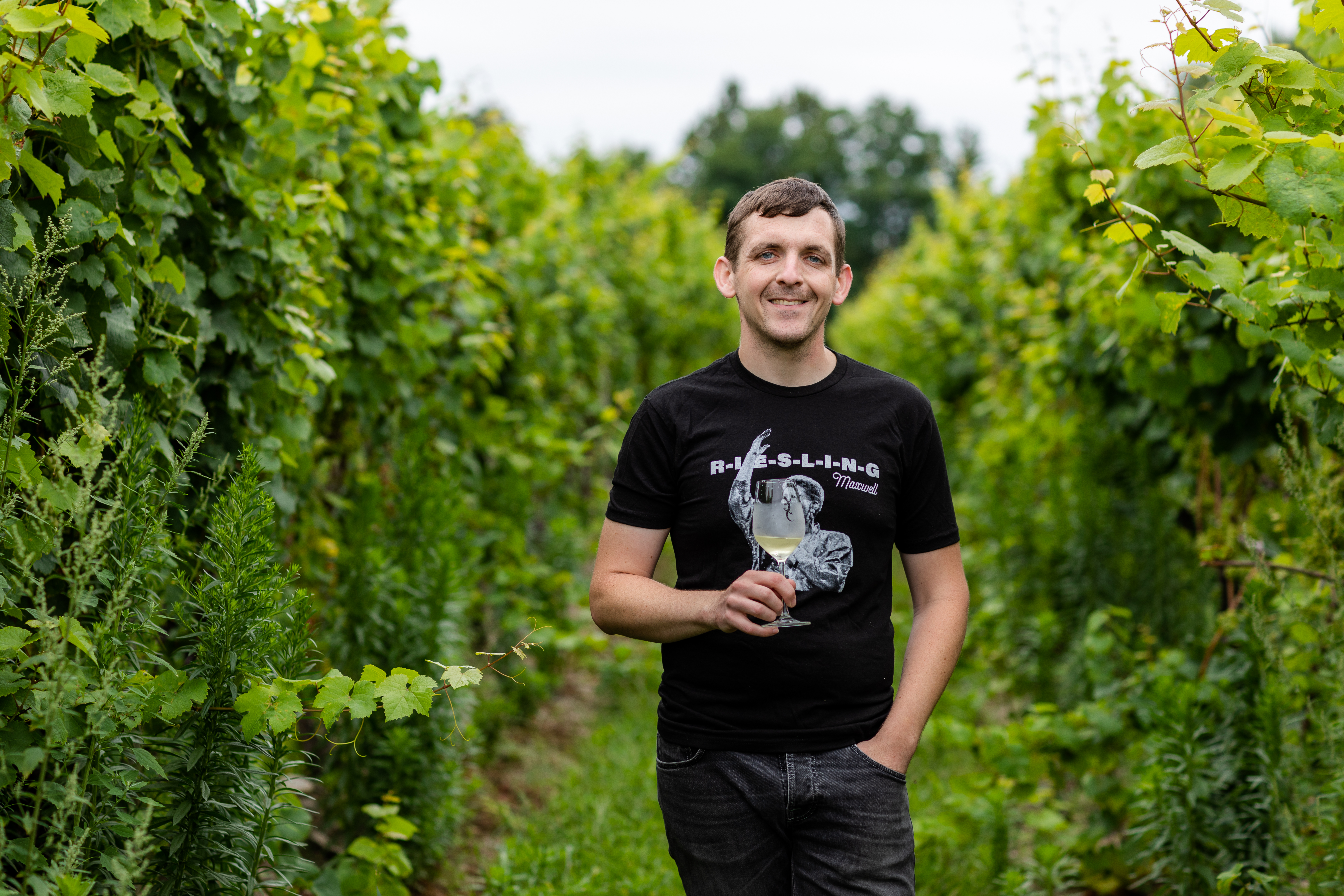 kelby james russell standing in a vineyard. he is wearing a black graphic tshirt and jeans, and holding a glass of white wine.